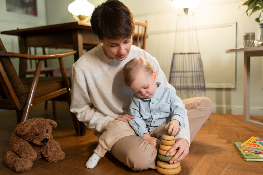 A babysitter is sitting on the floor with a toddler, helping him stack wooden rings. The scene is set in a cozy living room with wooden furniture, a soft toy bear, and some books on the floor. The babysitter is wearing a light-colored sweater and pants, while the toddler is dressed in a light blue shirt. Both appear focused and engaged in the activity.